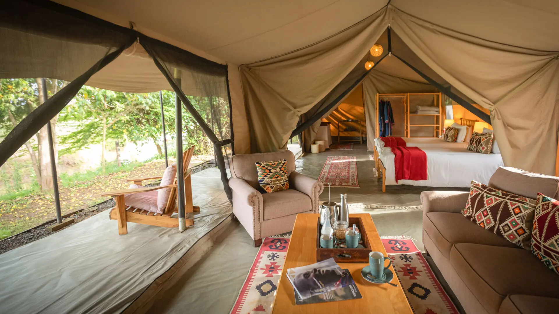 Interior of a large safari tent with seating area, beds, and open sides overlooking greenery