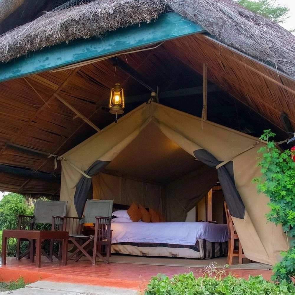 Luxury tent bed by chairs under a thatched roof with a lantern in Standard Tent Waterhole View at Serena Hotels & Resorts